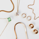 The LLUME Jewelry Vacation Capsule Set features thin gold chain necklaces—one with a green gemstone, one with a square clear stone—and pairs of gold hoop earrings, all arranged on a white surface for a sparkling display.