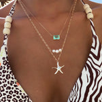 A person wears a zebra-print swimsuit with wooden beads, layered LLUME Jewelry Vacation Essentials Set gold necklaces featuring a starfish with green gemstone, three pearls, and a delicate pendant.