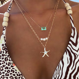 A person wears a zebra-print swimsuit with wooden beads, layered LLUME Jewelry Vacation Essentials Set gold necklaces featuring a starfish with green gemstone, three pearls, and a delicate pendant.