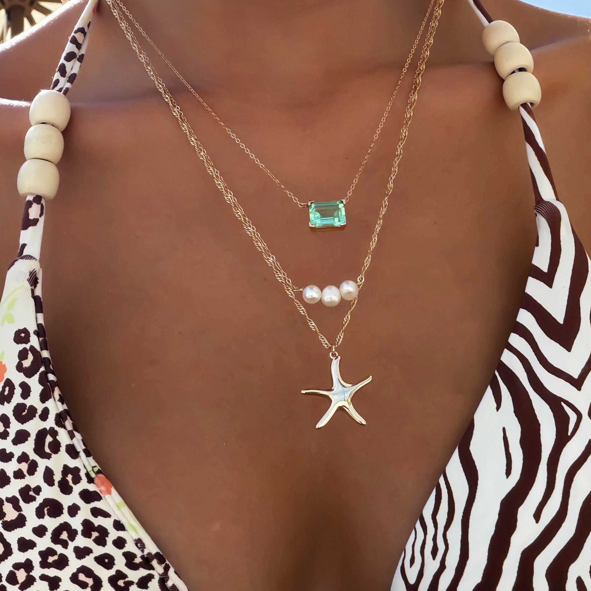 A person wears a zebra-print swimsuit with wooden beads, layered LLUME Jewelry Vacation Essentials Set gold necklaces featuring a starfish with green gemstone, three pearls, and a delicate pendant.
