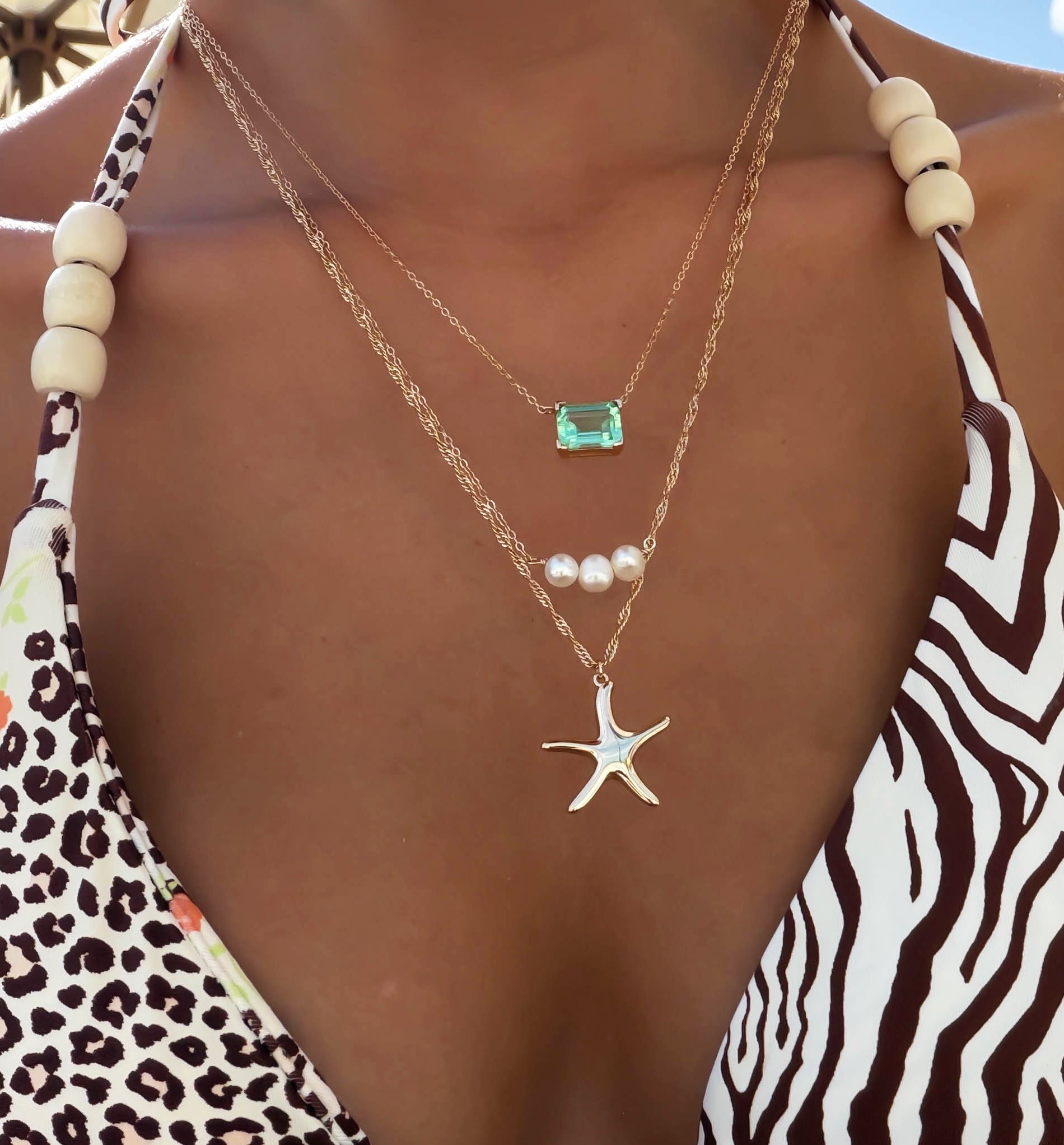 A person wears a zebra-print swimsuit with wooden beads, layered LLUME Jewelry Vacation Essentials Set gold necklaces featuring a starfish with green gemstone, three pearls, and a delicate pendant.