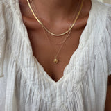 A person wearing a white textured blouse and the LLUME Jewelry Vintage Favorites Set—featuring three layered gold necklaces—shown from the shoulders to the chest.