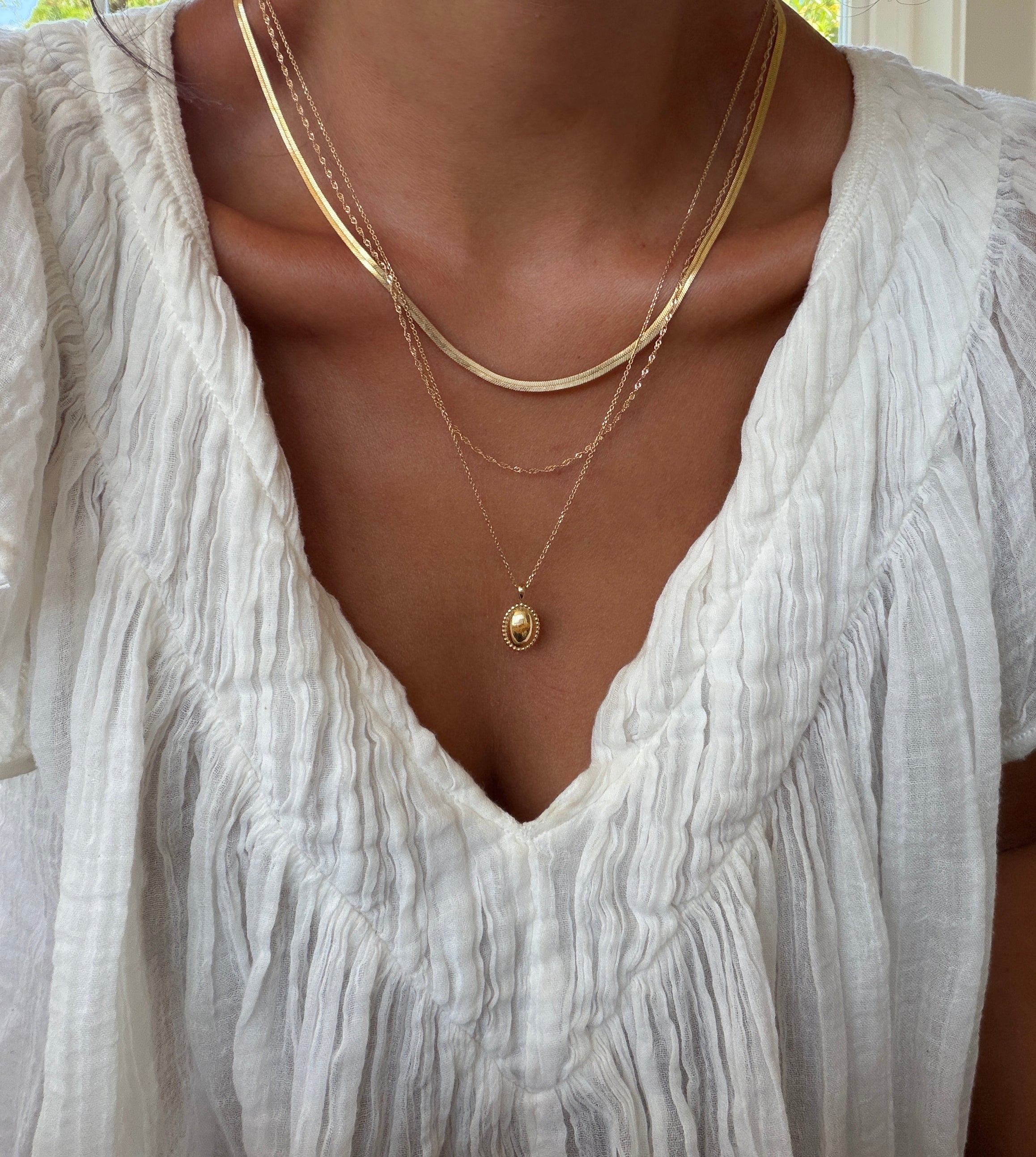 A person wearing a white textured blouse and the LLUME Jewelry Vintage Favorites Set—featuring three layered gold necklaces—shown from the shoulders to the chest.