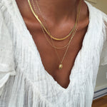 A person wearing a white textured top and three layered gold necklaces from the Vintage Favorites Set by LLUME Jewelry, with one featuring a small oval pendant, as natural light streams through a window in the background.