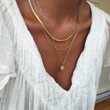 A person wearing a white textured top and three layered gold necklaces from the Vintage Favorites Set by LLUME Jewelry, with one featuring a small oval pendant, as natural light streams through a window in the background.