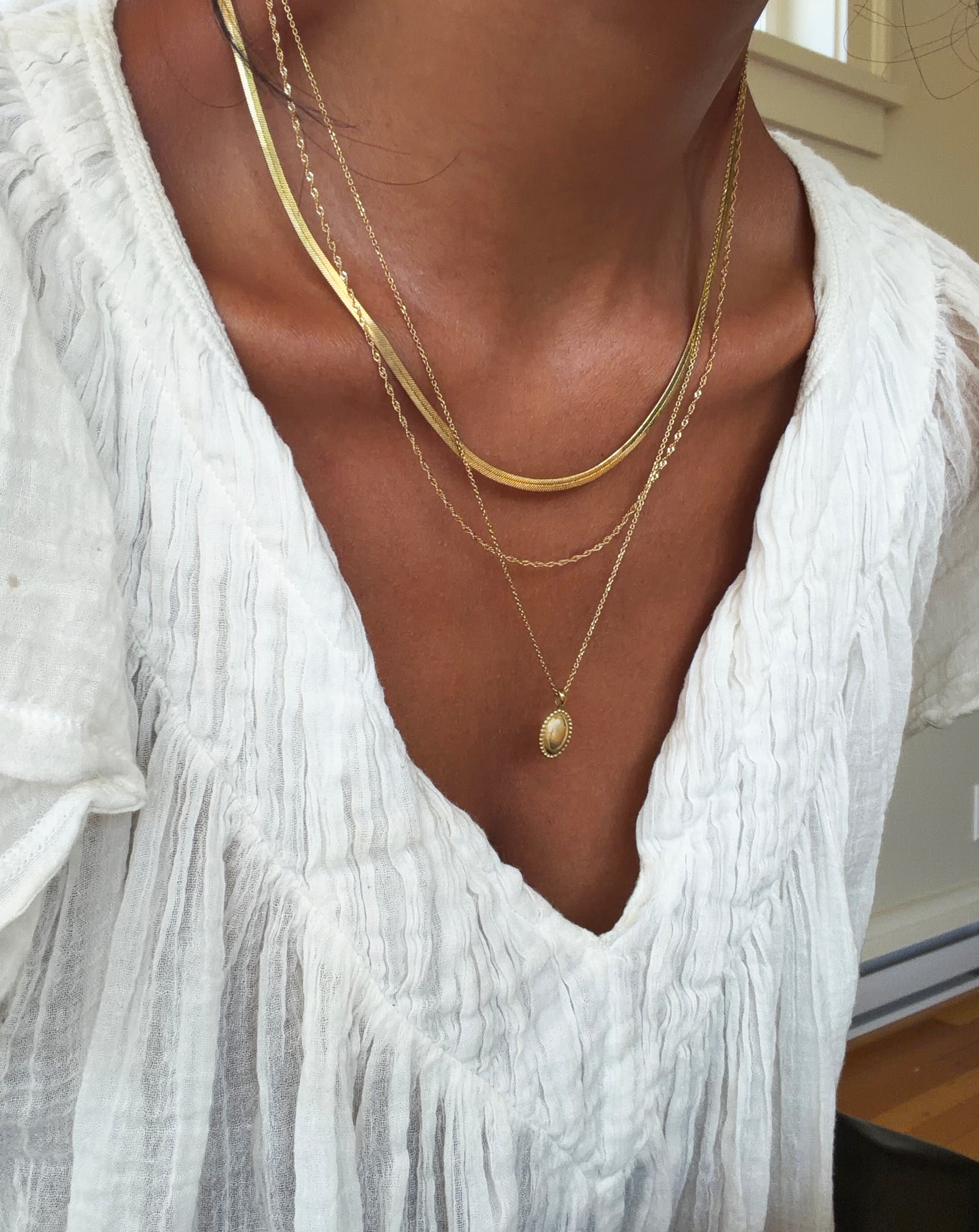 A person wearing a white textured top and three layered gold necklaces from the Vintage Favorites Set by LLUME Jewelry, with one featuring a small oval pendant, as natural light streams through a window in the background.
