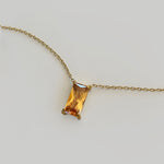 The Wild Honey Necklace by LLUME Jewelry features a rectangular, amber-colored gemstone pendant—reminiscent of honey—set in a simple gold prong setting, displayed against a light gray background.