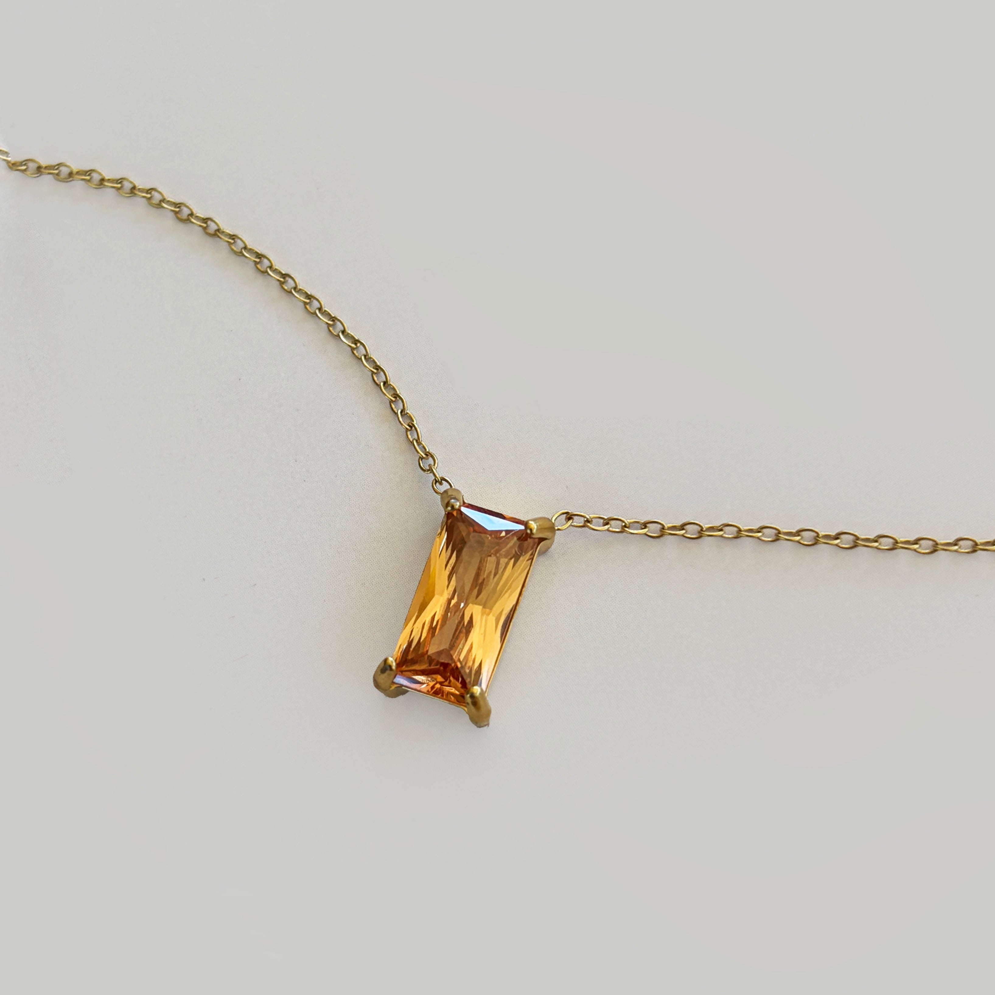 The Wild Honey Necklace by LLUME Jewelry features a rectangular, amber-colored gemstone pendant—reminiscent of honey—set in a simple gold prong setting, displayed against a light gray background.