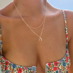 A person wearing a colorful floral dress and LLUME Jewelry’s Wishbone Daily Set necklace is shown from shoulders to mid-chest, sunlight illuminating their skin.